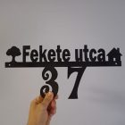 Kézben tartott fekete házikós házszámtábla Fekete utca 37 felirattal, a sín alatt lelógó házszámmal, szürke háttér előtt